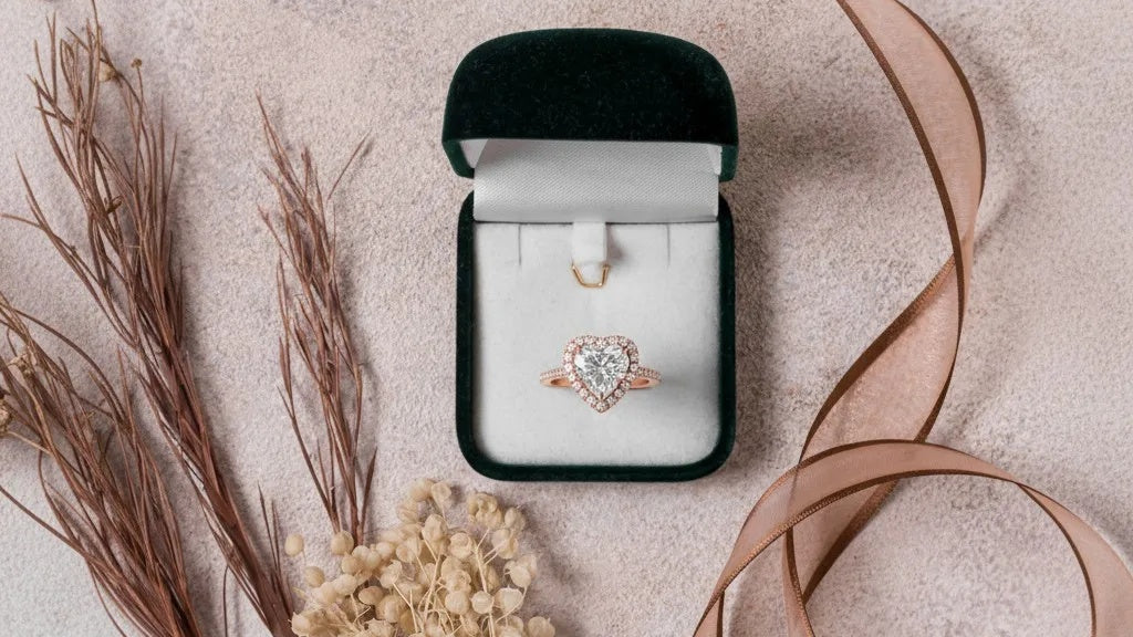 Heart-shaped diamond ring in a black velvet box on a beige surface with dried plants.